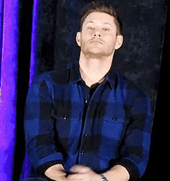 a man in a blue plaid shirt is sitting down with his hands folded .
