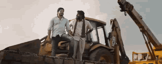 two men are standing next to a bulldozer on top of a brick wall .