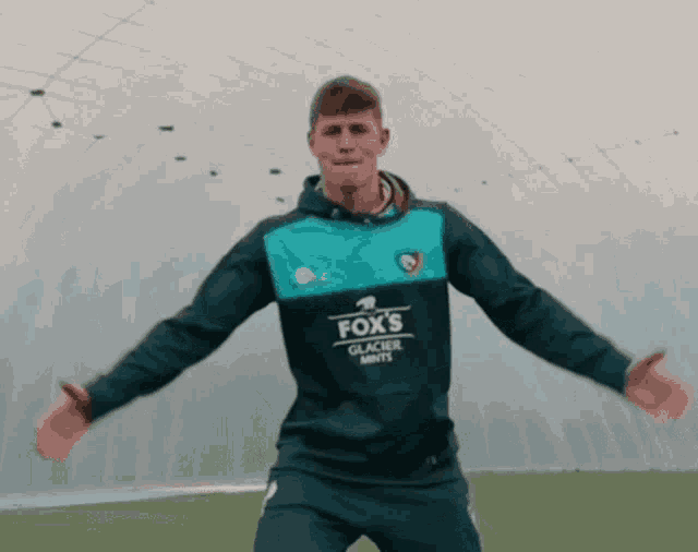 a man wearing a fox 's glacier mints sweatshirt is standing with his arms outstretched