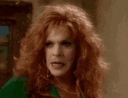 a woman with red hair and a green shirt is wearing a wig and making a funny face .