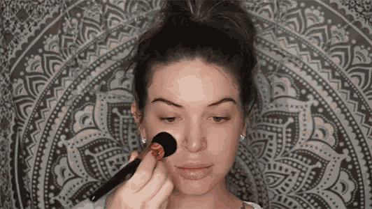 a woman is applying makeup to her face with a brush in front of a mandala tapestry .