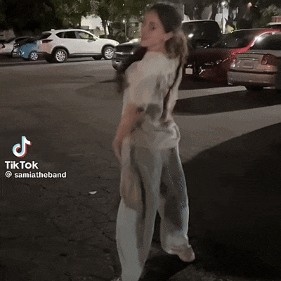 a woman in a tie dye shirt is standing in front of a parking lot with cars parked on the side of the road
