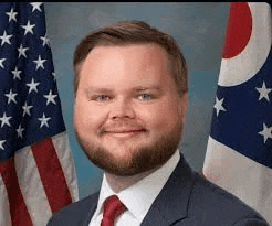 a man with a beard is wearing a suit and tie and smiling in front of an american flag .