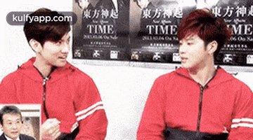 two young men in red jackets are standing next to each other in front of a wall with posters for time .