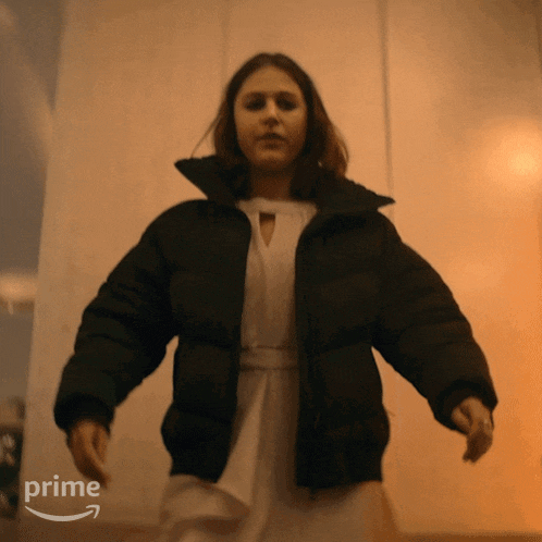 a woman wearing a black jacket and a white dress is dancing in front of a wall with the word prime on it