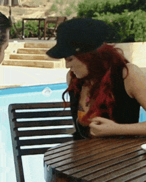 a woman wearing a black hat sits at a table by a pool