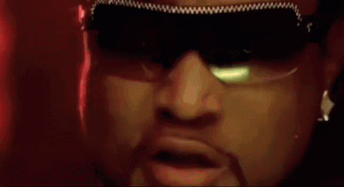 a man wearing sunglasses and a necklace is standing in front of a crowd of people in a club .