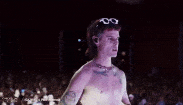 a man without a shirt is standing in front of a crowd wearing sunglasses .