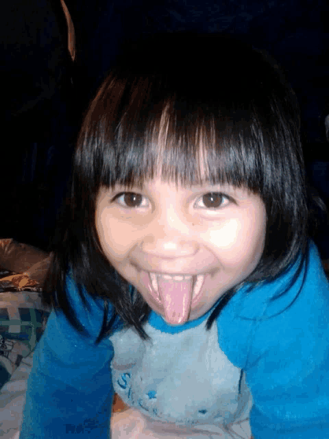 a little girl sticking out her tongue and wearing a blue shirt that says s on it