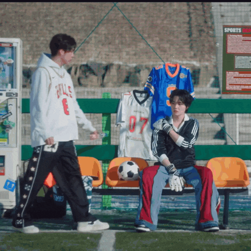 a man wearing a sweatshirt that says bulls on it walks past a man sitting on a bench with a soccer ball