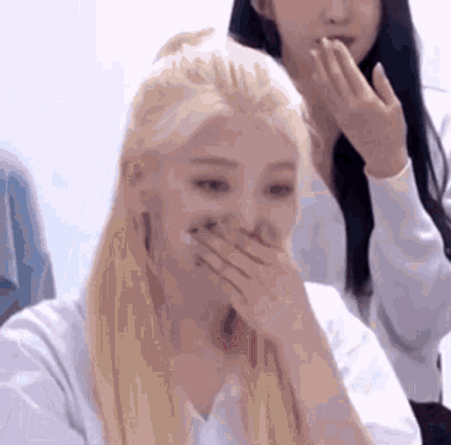 a woman with blonde hair is covering her mouth with her hands while another woman looks on .