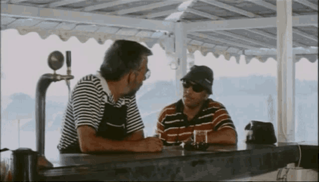 two men sitting at a bar with one wearing sunglasses and the other wearing a hat