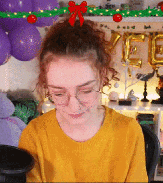 a woman wearing glasses and a yellow sweater stands in front of balloons and a christmas tree