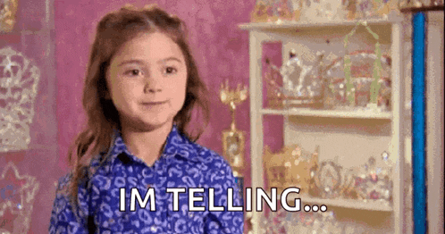 a little girl in a blue shirt is standing in front of a shelf with trophies and says `` im telling ... '' .