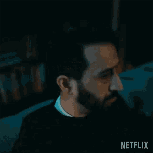 a man with a beard is sitting on a couch in a dark room looking at something .
