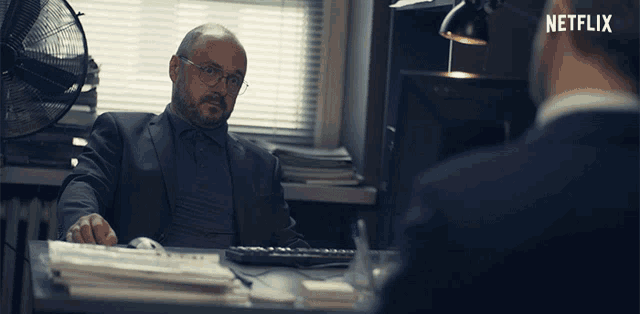 a man in a suit sits at a desk with a netflix logo in the background