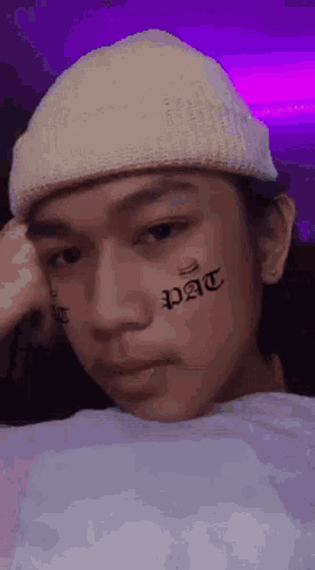 a young man with a tattoo on his face is wearing a beanie and a white shirt .