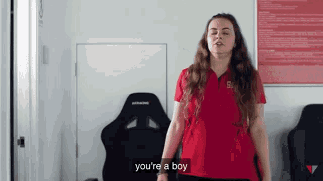 a woman in a red shirt is standing in front of a black chair and says you 're a boy