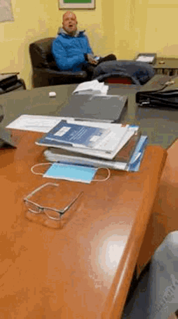 a man in a blue jacket is sitting at a desk with a stack of papers and a mask on it .