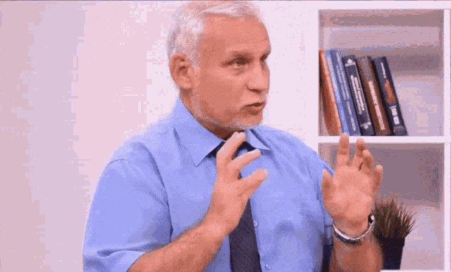 a man in a blue shirt and tie is talking in front of a bookshelf with books on it