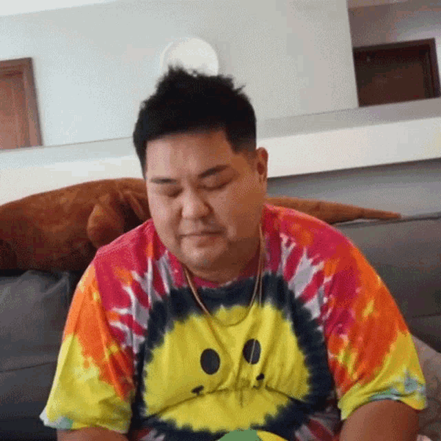 a man is sitting on a couch wearing a tie dye shirt and a necklace .