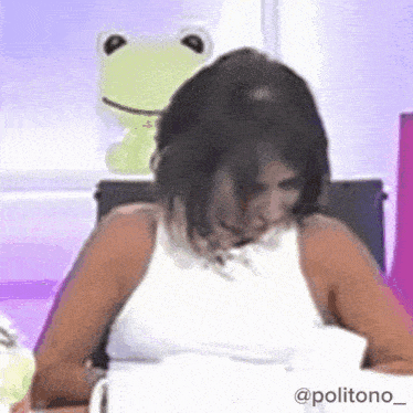 a woman in a white tank top is sitting at a table with a stuffed frog behind her .