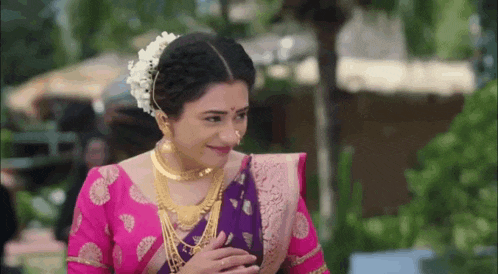 a woman in a pink and purple saree and gold jewelry is smiling and looking at the camera .