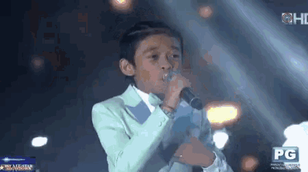 a young boy singing into a microphone on a stage with a pg logo behind him