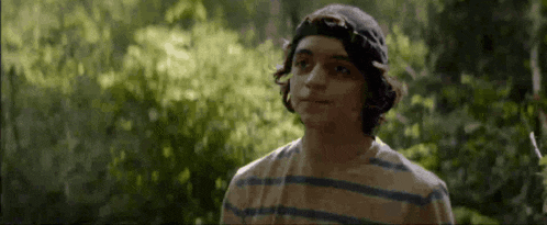a young man wearing a hat and striped shirt is standing in the woods .