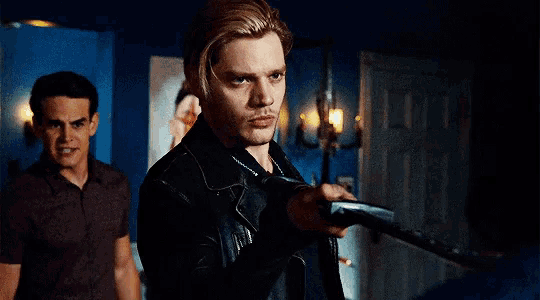 a man in a leather jacket is holding a gun while another man stands behind him .