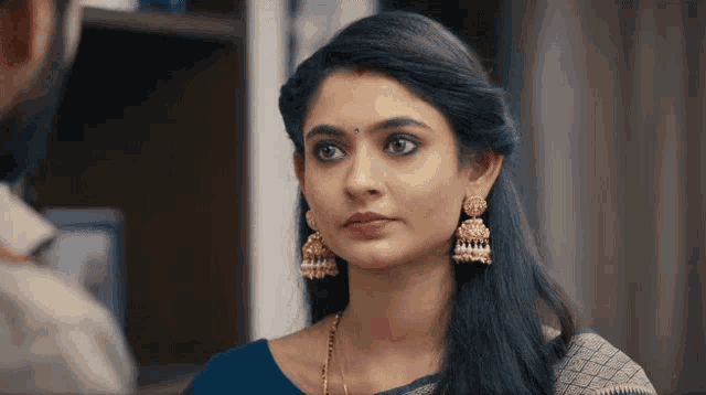 a woman wearing earrings and a blue blouse is looking at a man in a room .
