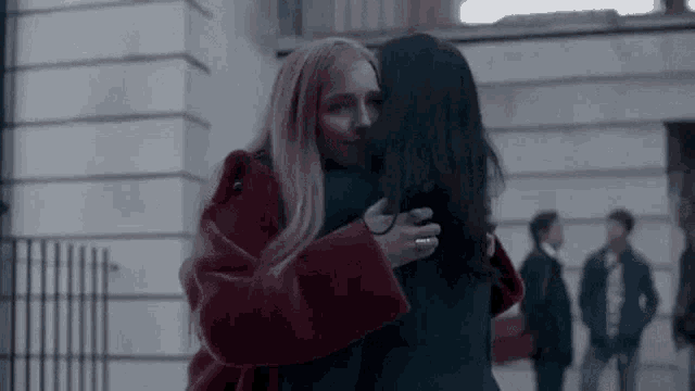 two women are hugging each other in front of a building . one of the women is wearing a red coat .