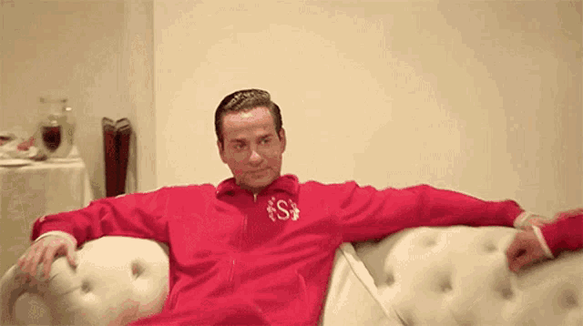 a man is sitting on a couch wearing a red jacket with the letter s on it