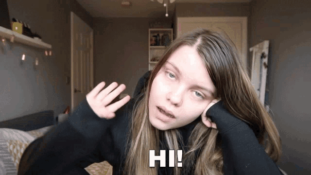 a girl in a black sweater says hi with her hand on her face