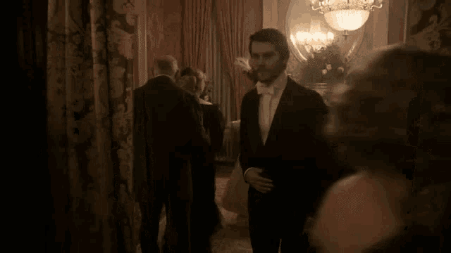 a man in a tuxedo and bow tie is standing in a dark room with other people .