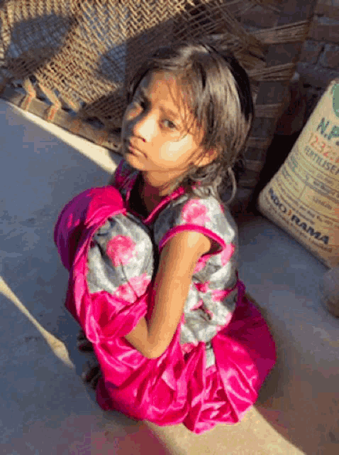 a little girl in a pink dress sits on the ground next to a bag of np fertilizer