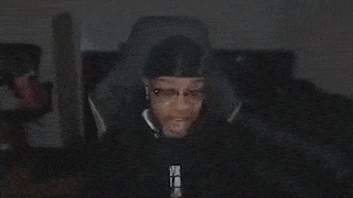 a man wearing a hat and glasses is making a funny face while sitting in a dark room .