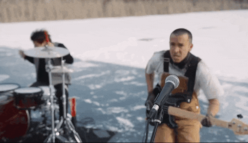 a man is playing a guitar in front of a microphone while another man plays drums behind him .