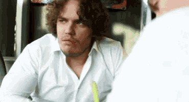 a man with curly hair and a beard is sitting at a table talking to another man .