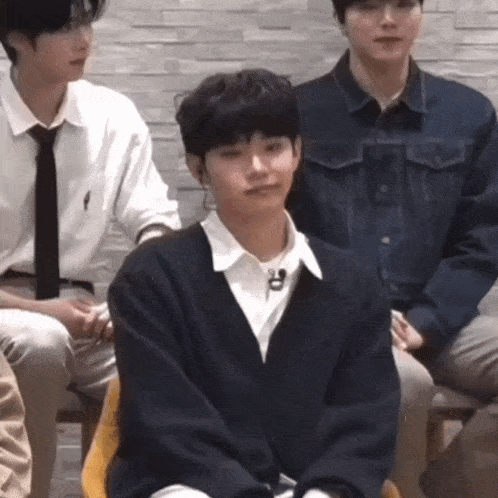 a group of young men are sitting in a room with one wearing a black sweater and tie