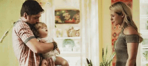 a man is holding a baby in his arms while a woman looks on in a living room .