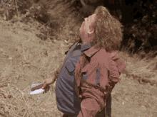 a man in a red shirt is standing on a dirt road looking up at the sky .