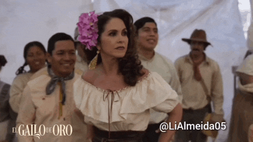 a woman with a flower in her hair is standing in front of a group of people with the caption el gallo de oro
