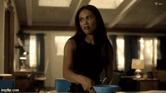 a woman in a black tank top is stirring a blue bowl on a table with imgflip.com in the corner