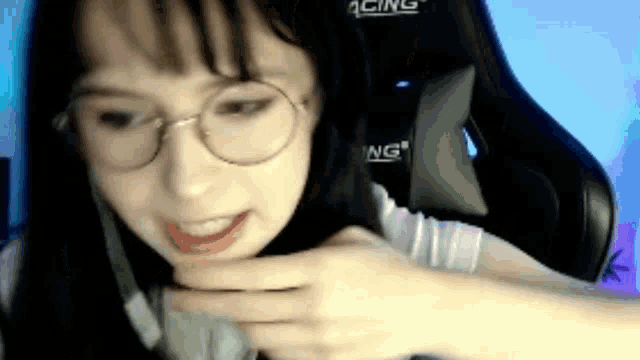 a woman wearing glasses is sitting in a gaming chair and making a funny face .