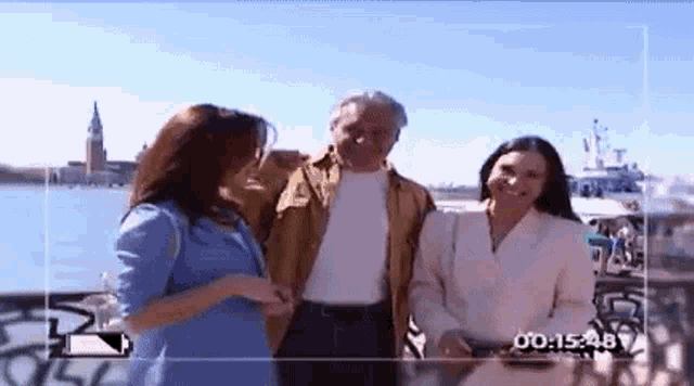 a man and two women are standing next to each other in front of a body of water with a time stamp of 0 : 15.48