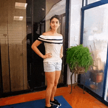 a woman in shorts and a off the shoulder top stands in front of a window