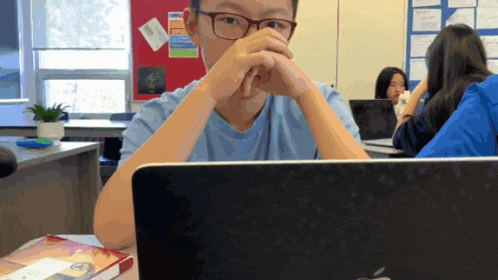a boy wearing glasses sits in front of a laptop in a classroom