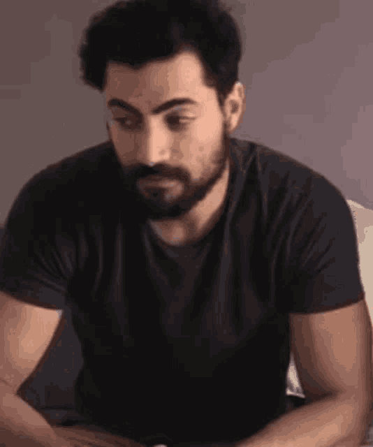 a man with a beard wearing a black shirt is sitting on a bed .