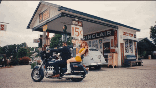 a man and a woman are riding a motorcycle in front of a gas station that says sinclair